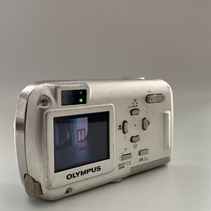 May include: A silver Olympus digital camera with a black and white LCD screen displaying "NO  TARGET" and a red exclamation point. The camera has a black button labeled "OK" and a silver button labeled "QUICK VIEW".