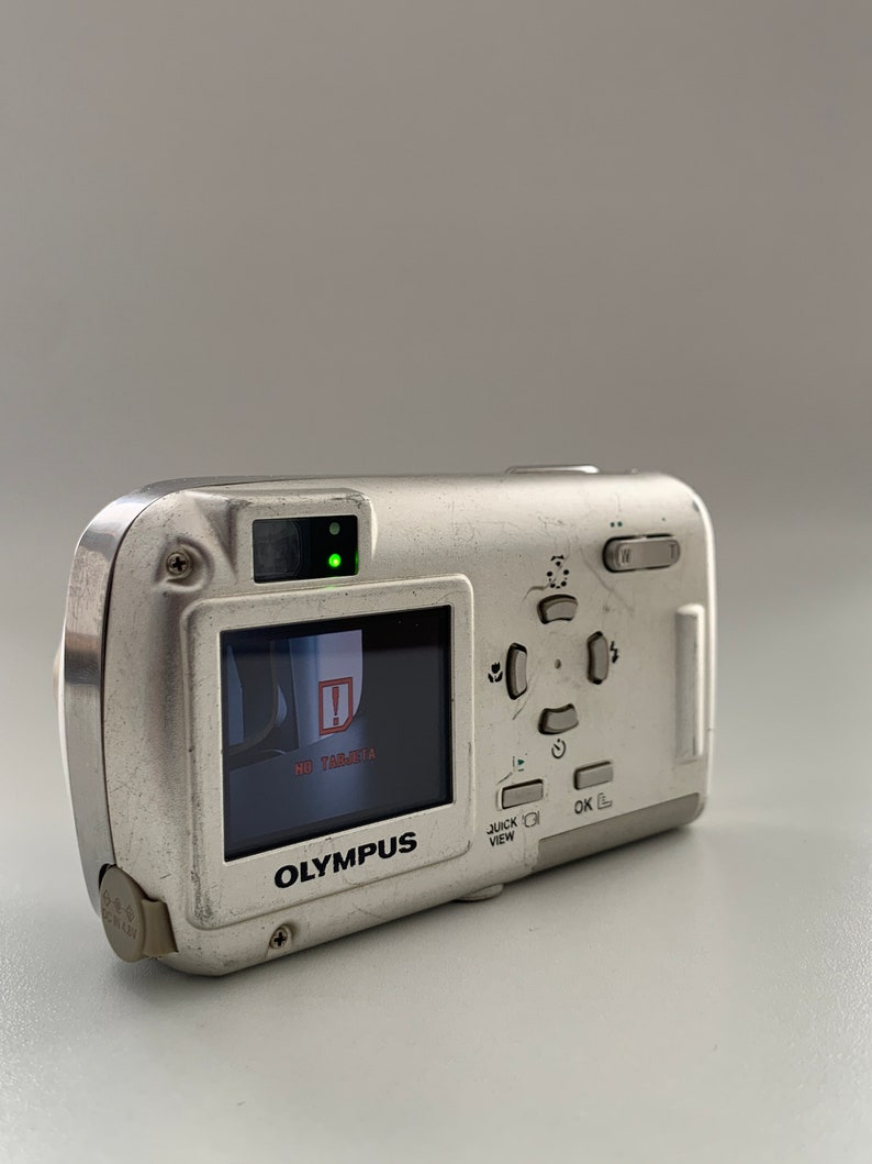 May include: A silver Olympus digital camera with a black and white LCD screen displaying "NO  TARGET" and a red exclamation point. The camera has a black button labeled "OK" and a silver button labeled "QUICK VIEW".