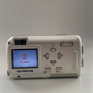 May include: A silver Olympus digital camera with a black and white screen that reads "NO TARJETA". The camera has a variety of buttons, including a quick view button, an OK button, and a directional pad.