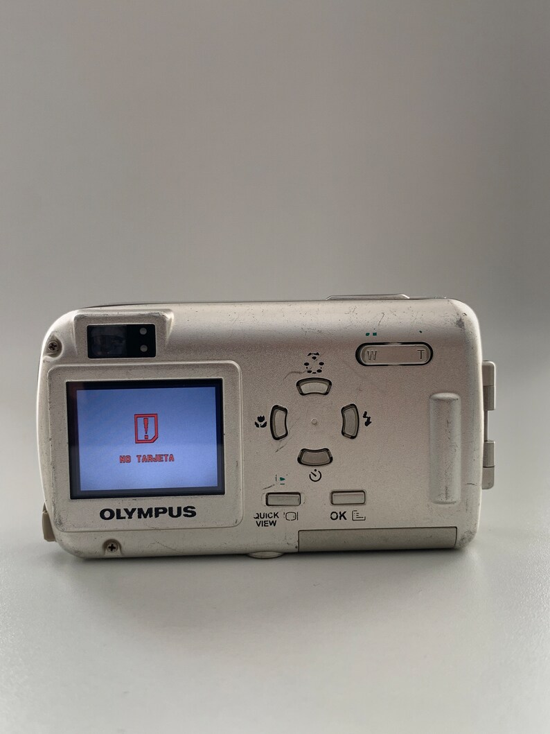 May include: A silver Olympus digital camera with a black and white screen that reads "NO TARJETA". The camera has a variety of buttons, including a quick view button, an OK button, and a directional pad.