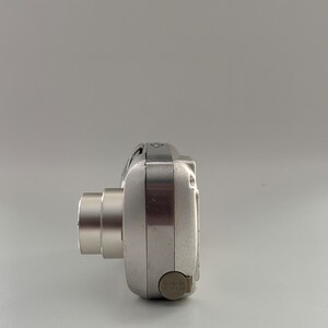 May include: A silver digital camera with a lens on the left side and a DC IN 6.8V label on the right side.