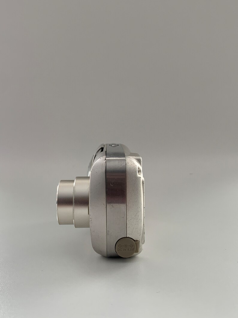 May include: A silver digital camera with a lens on the left side and a DC IN 6.8V label on the right side.