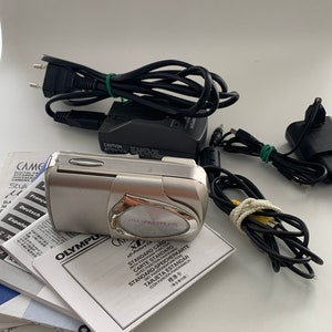 May include: A silver Olympus digital camera with a black lens and a black and silver charger. The camera is sitting on a stack of instruction manuals in multiple languages. The manuals include instructions for using the camera and for inserting an xD-Picture Card.