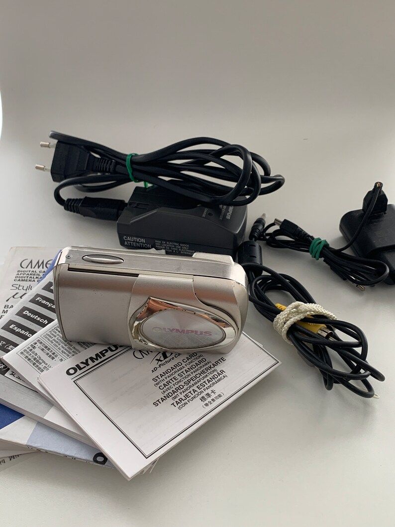 May include: A silver Olympus digital camera with a black lens and a black and silver charger. The camera is sitting on a stack of instruction manuals in multiple languages. The manuals include instructions for using the camera and for inserting an xD-Picture Card.