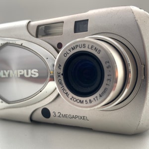 May include: A silver Olympus digital camera with a 3.2 megapixel resolution and a 3x optical zoom lens. The lens has the text "Olympus Lens AF 3x Optical Zoom 5.8-17.4mm 1:3.1-5.2" printed on it.