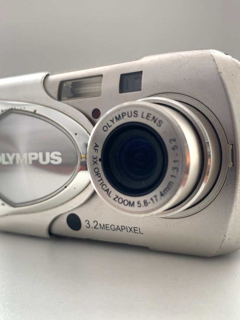 May include: A silver Olympus digital camera with a 3.2 megapixel resolution and a 3x optical zoom lens. The lens has the text "Olympus Lens AF 3x Optical Zoom 5.8-17.4mm 1:3.1-5.2" printed on it.