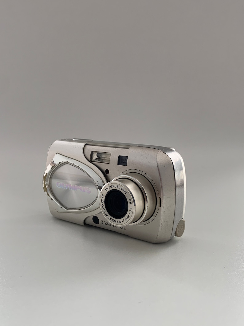 May include: A silver Olympus digital camera with a 3.2 megapixel lens and a 3x optical zoom. The camera has a silver body with a black lens.