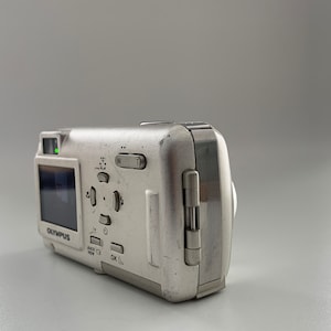 May include: A silver Olympus digital camera with a small LCD screen and buttons for zoom, playback, and menu options. The camera has a built-in flash and a lens cover.