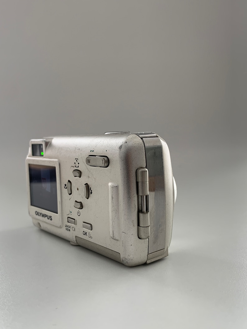 May include: A silver Olympus digital camera with a small LCD screen and buttons for zoom, playback, and menu options. The camera has a built-in flash and a lens cover.