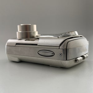 May include: A silver digital camera with a lens cover and a small LCD screen on the back. The camera has a sleek, modern design.