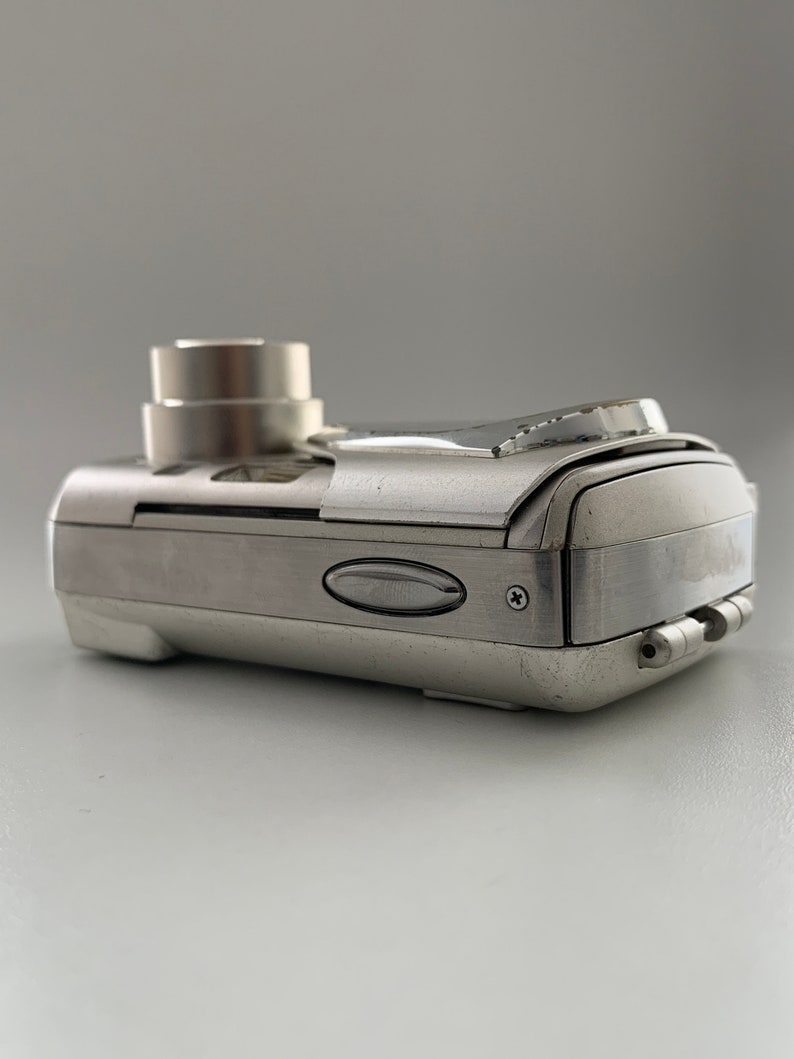 May include: A silver digital camera with a lens cover and a small LCD screen on the back. The camera has a sleek, modern design.