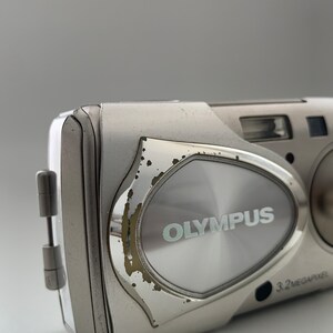 May include: A silver Olympus digital camera with a 3.2 megapixel resolution. The camera has a silver metal body with a slightly worn finish.