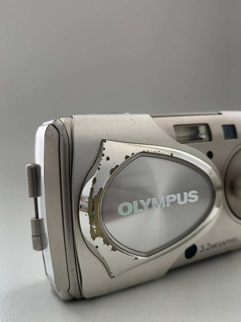 May include: A silver Olympus digital camera with a 3.2 megapixel resolution. The camera has a silver metal body with a slightly worn finish.