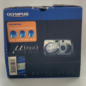 May include: A blue box with a silver Olympus M[mju:] digital camera on the front. The box has the text "Olympus" and "M[mju:] 300 Digital" on it. The camera has a silver metal body and a black lens.