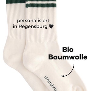 May include: Pair of white crew socks with two green stripes. The socks are made of organic cotton and are personalized with the text "personalisiert in Regensburg" and a black heart. The socks are made in Portugal and are a product of the brand "plattohclock".