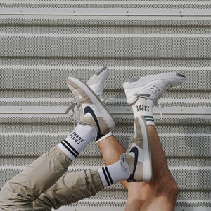May include: Two pairs of white sneakers with black accents and white socks with black text that reads "SOCIAL" on them. The sneakers are held up in the air against a gray metal background.