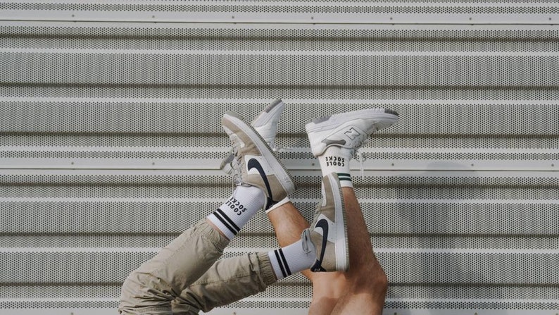 May include: Two pairs of white sneakers with black accents and white socks with black text that reads "SOCIAL" on them. The sneakers are held up in the air against a gray metal background.