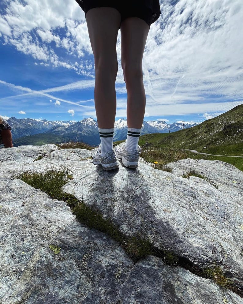 May include: A person wearing white socks with black stripes and white sneakers stands on a rocky surface with a mountain range in the background. The sky is blue with white clouds.