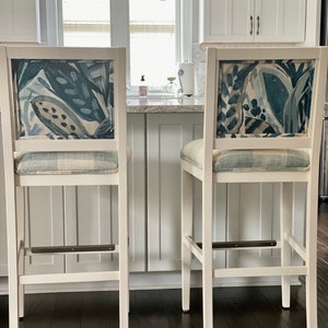 May include: Two white bar stools with a blue and white patterned fabric seat back and a white fabric seat. The stools are on a dark wood floor.