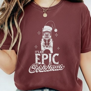 May include: Burgundy t-shirt with a white graphic featuring a tower wearing a Santa hat, surrounded by stars, and the text "It's an EPIC Christmas" in a distressed font. The shirt is a solid color.