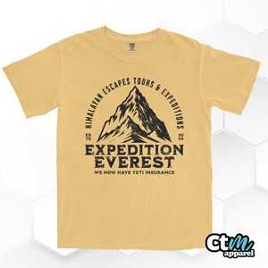 Op de afbeelding: Mosterdgeel t-shirt met een zwarte, verweerde afbeelding. De afbeelding toont een bergsilhouette met de tekst "Himalayan Escapes Tours & Expeditions" en "Expedition Everest". Het shirt bevat ook de tekst "We Now Have Yeti Insurance".