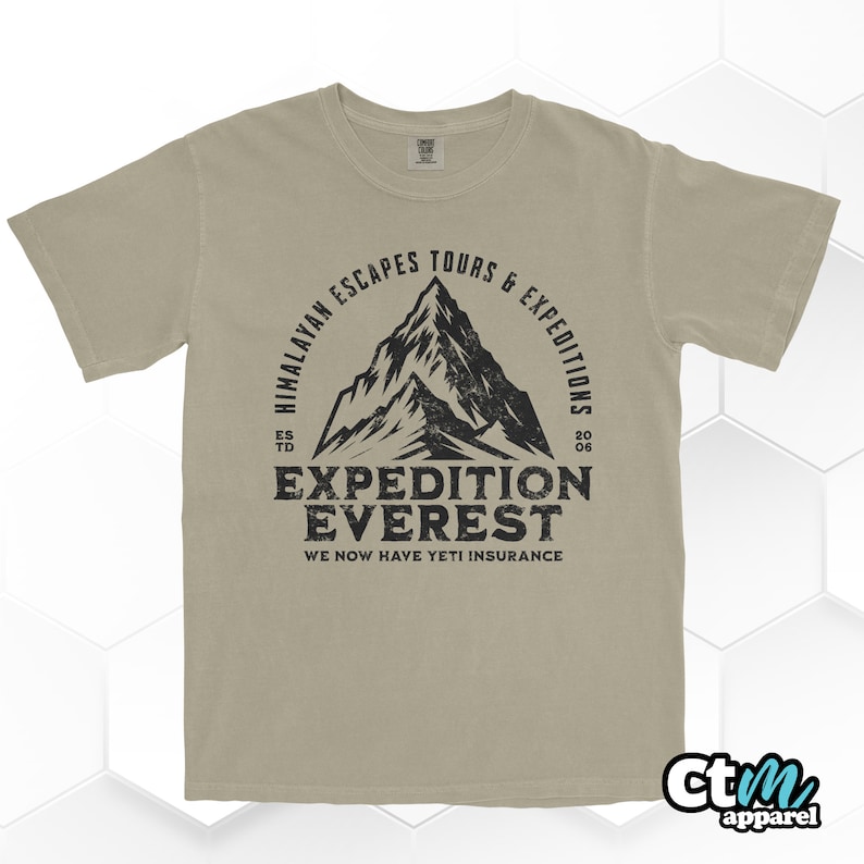 Op de afbeelding: Een beige t-shirt met een zwarte grafische print. Het ontwerp bevat een bergillustratie en de tekst "Himalayan Escapes Tours & Expeditions" en "Expedition Everest". De tekst "We Now Have Yeti Insurance" is ook inbegrepen.
