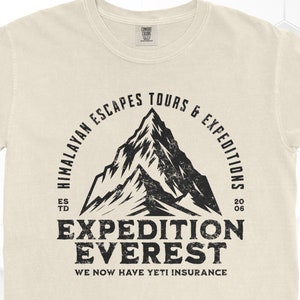 Op de afbeelding: Cr&egrave;mekleurig T-shirt met een zwarte bergafbeelding in distress-stijl en de woorden "EXPEDITION EVEREST" en "HIMALAYAN ESCAPES TOURS & EXPEDITIONS". De tekst "WE NOW HAVE YETI INSURANCE" is ook opgenomen.