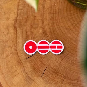 May include: A red and white sticker featuring three connected circles. The first circle has a white dot, the second has horizontal lines, and the third has a horizontal line and a vertical line. The sticker is on a wooden surface.
