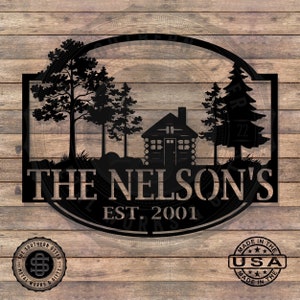 May include: Black metal silhouette of a cabin in the woods with two trees on either side. The text "THE NELSON'S EST. 2001" is below the cabin.