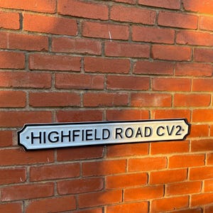 May include: A white sign with black lettering on a brick wall. The sign reads "HIGHFIELD ROAD CV2".