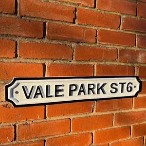 May include: White and black street sign with rounded edges and black lettering that reads "VALE PARK ST6". The sign is mounted on a brick wall.