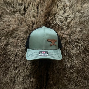 May include: A green and black trucker hat with a brown leather patch featuring an anvil and the text "Anvil Time". The hat is made by Richardson 112.