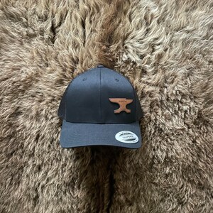May include: A black mesh trucker hat with a brown leather anvil logo. The hat has a black brim and a black mesh back.