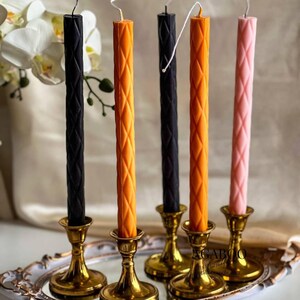 May include: Five tall, slender taper candles with a diamond-patterned texture, featuring two black, two orange, and one light pink candle. Each candle is held in a polished gold-toned metal candlestick holder, all arranged on a decorative tray.