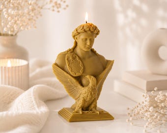May include: A yellow beeswax candle shaped as a bust of a classical figure wearing a laurel wreath, with an eagle perched on the chest. The figure has a shield-like emblem on the shoulder and rests on a square base. A single lit wick burns at the top.
