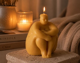 May include: A yellow beeswax candle shaped like a seated figure with knees pulled towards the chest. The candle is lit, with a small flame at the top of the head. The figure has a smooth, rounded form and is positioned on a textured stone pedestal.