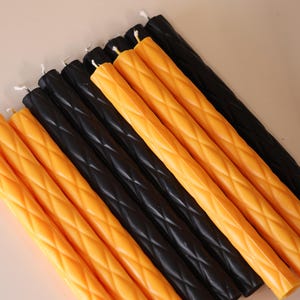 May include: A collection of ten long, cylindrical taper candles arranged in a parallel row. Five candles are a vibrant orange color and five are black. Each candle features a raised, diamond-shaped lattice texture along its entire length.