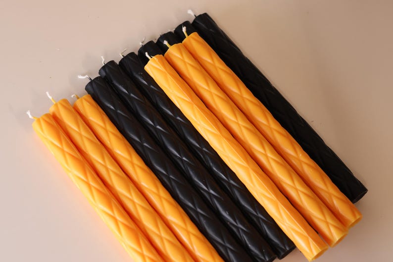 May include: A collection of ten long, cylindrical taper candles arranged in a parallel row. Five candles are a vibrant orange color and five are black. Each candle features a raised, diamond-shaped lattice texture along its entire length.