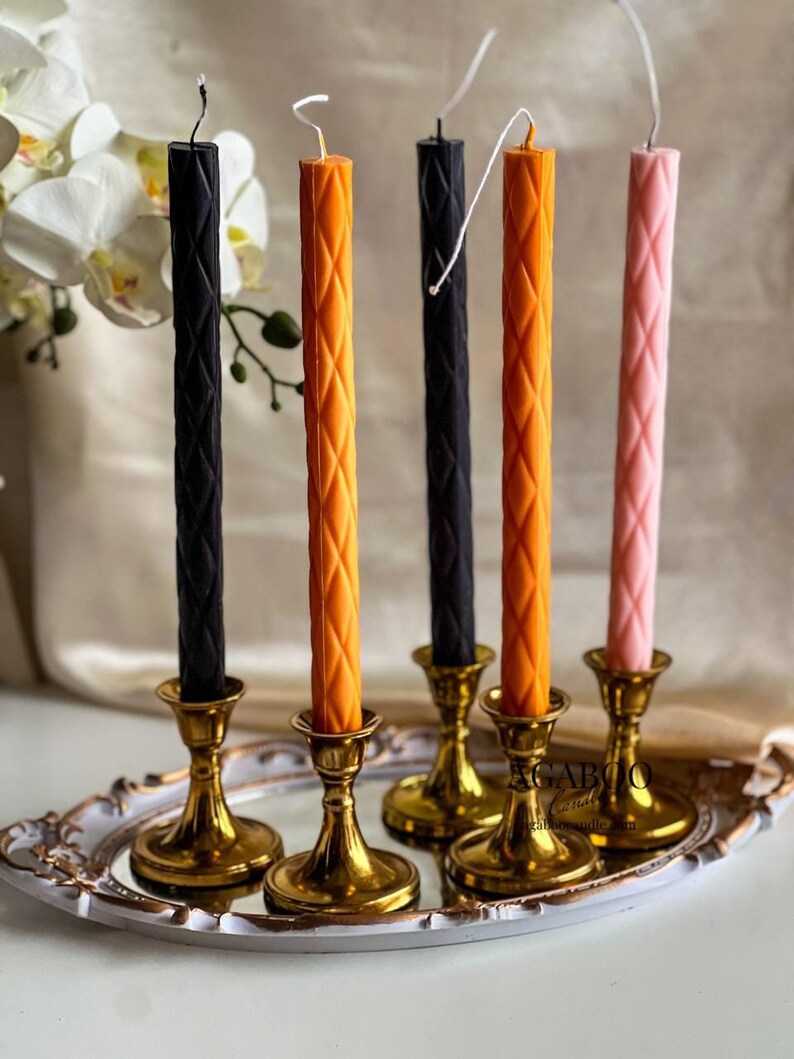 May include: Five tall, slender taper candles with a diamond-patterned texture, featuring two black, two orange, and one light pink candle. Each candle is held in a polished gold-toned metal candlestick holder, all arranged on a decorative tray.