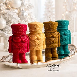 May include: Four nutcracker-shaped candles in red, light yellow, brown, and teal. Each candle features a detailed uniform, a mustache, and a hat with a shell emblem. The text AGABOO Candles and agaboocandle.com is printed on the silver tray.