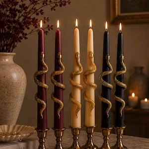 May include: Six tall taper candles in burgundy, cream, and black, each wrapped with a textured gold-coloured snake ornament. The candles are lit and stand upright in matching metallic gold candle holders arranged in a row on a flat surface.