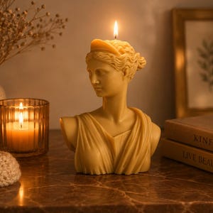 May include: A light yellow beeswax candle shaped like a classical bust of a person with a headband, featuring a lit wick at the top. The sculpture depicts a figure with draped clothing over the shoulders, rendered in a smooth, solid wax material.