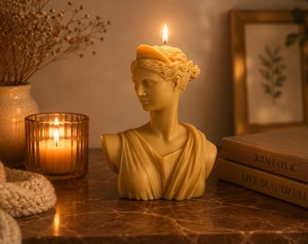 Op de afbeelding: Een lichtgele bijenwaskaars in de vorm van een klassieke buste van een persoon met een hoofdband, voorzien van een brandende lont aan de bovenkant. Het beeld toont een figuur met gedrapeerde kleding over de schouders, gemaakt van gladde was.