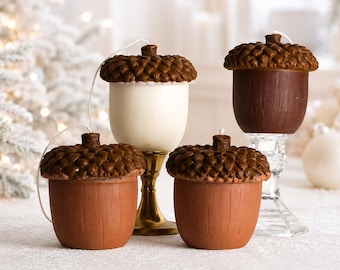 May include: Four acorn-shaped candles with textured brown caps. Two candles have terracotta-colored bases, one has a dark brown base, and one has a cream-colored base. The candles are displayed on gold and clear glass pedestals with white wicks.
