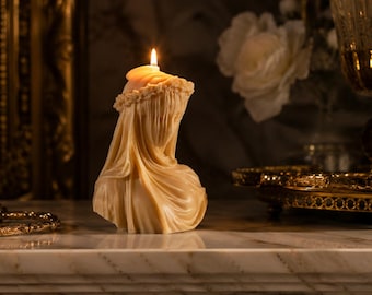 May include: A cream-coloured wax candle sculpted into the bust of a figure wearing a draped veil and a floral crown. The candle is lit, with a small flame burning at the top of the head. The sculpture features intricate folds in the fabric.