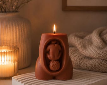 May include: A terracotta-colored pillar candle shaped like a human torso with a central cutout featuring a sculpted figure in a fetal position. The candle has a single lit wick at the top, casting a warm glow across its smooth, matte surface.