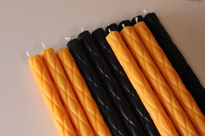 May include: A collection of long, slender taper candles arranged in a row. The candles feature a repeating diamond-shaped embossed pattern along their length and are colored in alternating sections of vibrant orange and solid black wax.
