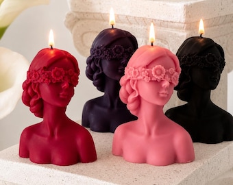 Puede incluir: Cuatro velas con forma de busto en rojo, morado oscuro, rosa y negro. Cada vela representa a una persona con una corona de flores cubriendo los ojos y una mecha encendida en la parte superior. Están dispuestas sobre una superficie de piedra.