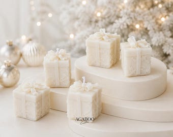 May include: Five square off-white candles shaped like gift boxes with embossed lace patterns and molded bows on top. Each candle features a central wick. The text AGABOO Candles is printed in black on the base of the front-most candle.