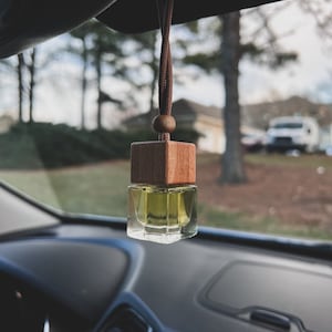 May include: A brown wooden car air freshener with a clear glass bottle filled with a yellow liquid. The air freshener is hanging from the rearview mirror of a car.
